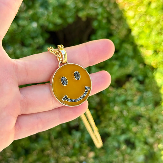 Hand holding a gold necklace with a smiley face pendant against a blurred green background