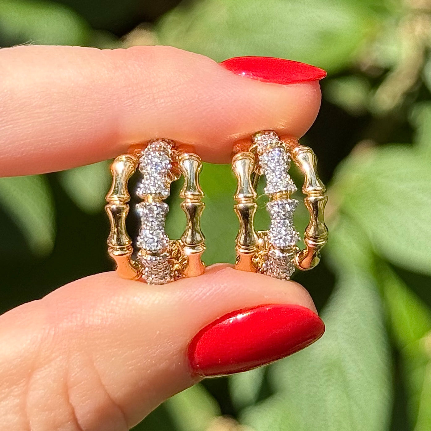 Gold and diamond earrings on a finger with red nail polish against a green leafy background