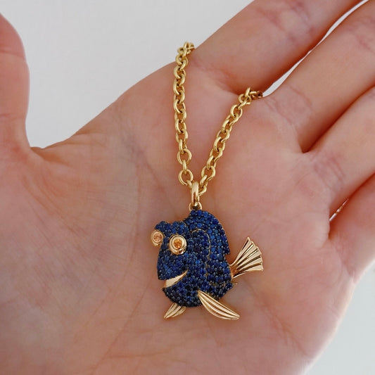 Gold and blue fish-shaped pendant on a chain held in a hand against a white background
