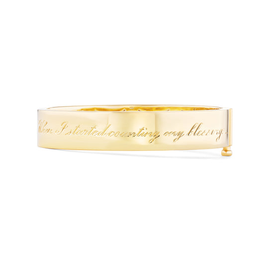 A gold bangle bracelet with hand engraving on it, displayed against a white background.