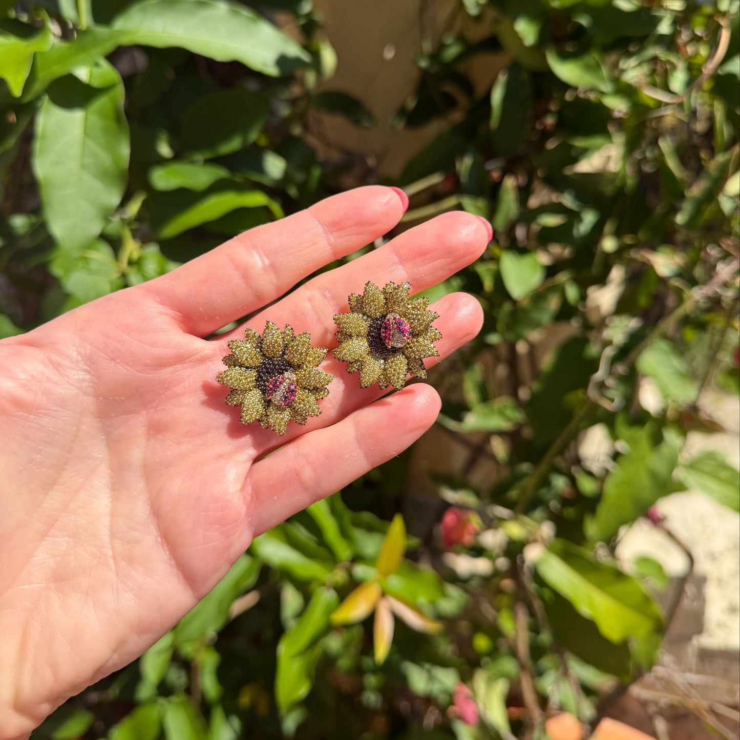 Hand holding two sunflower earrings with ladybugs on a blurred green background