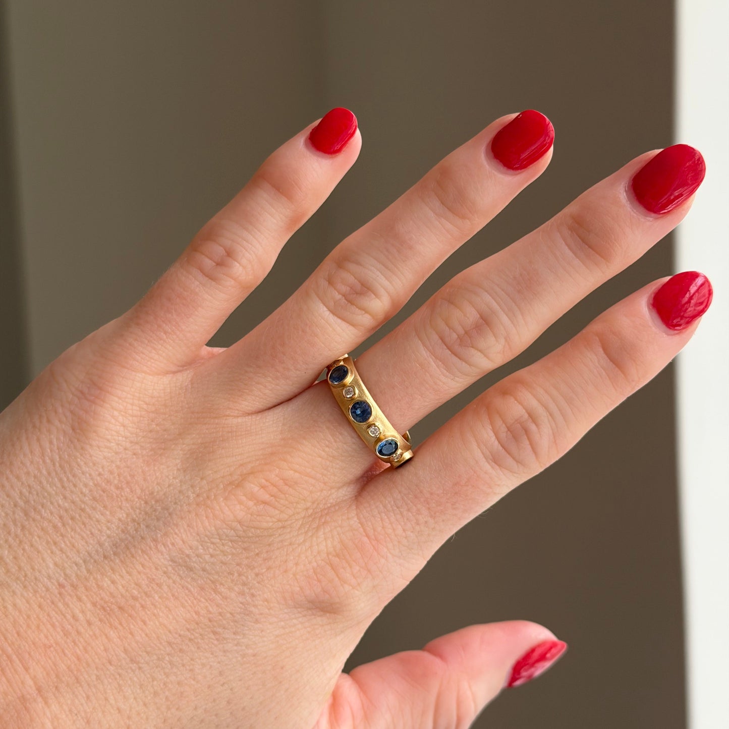 Hand wearing a gold ring with colored stones against a neutral background