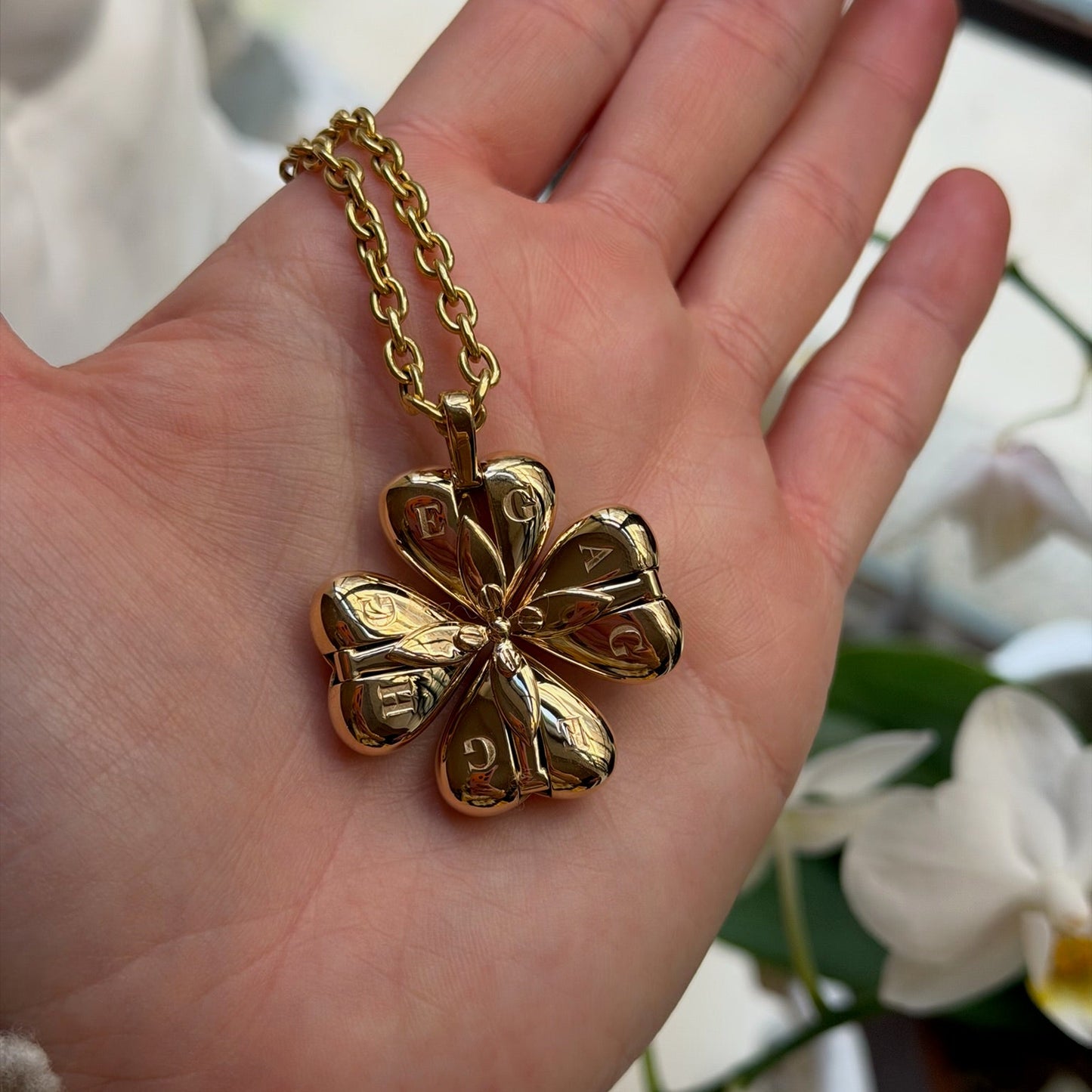 Gold four-leaf clover pendant on a chain held in a hand with a blurred floral background