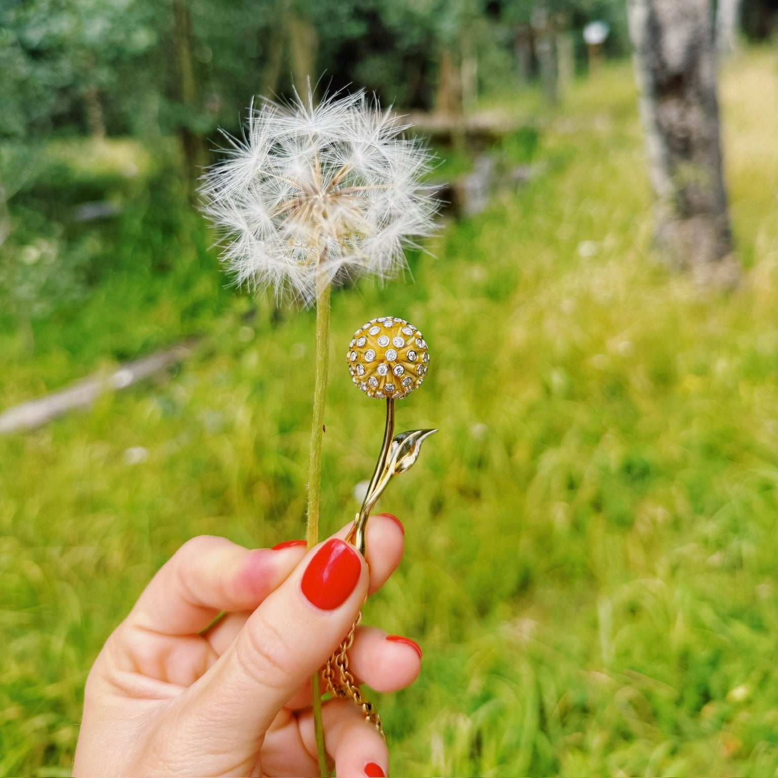 Hand holding a dandelion with a decorative gold stem against a blurred natural background