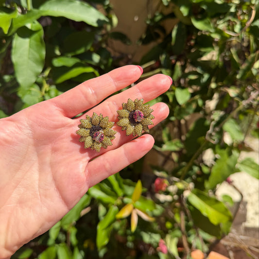 Hand holding two sunflower earrings with ladybugs on a blurred green background